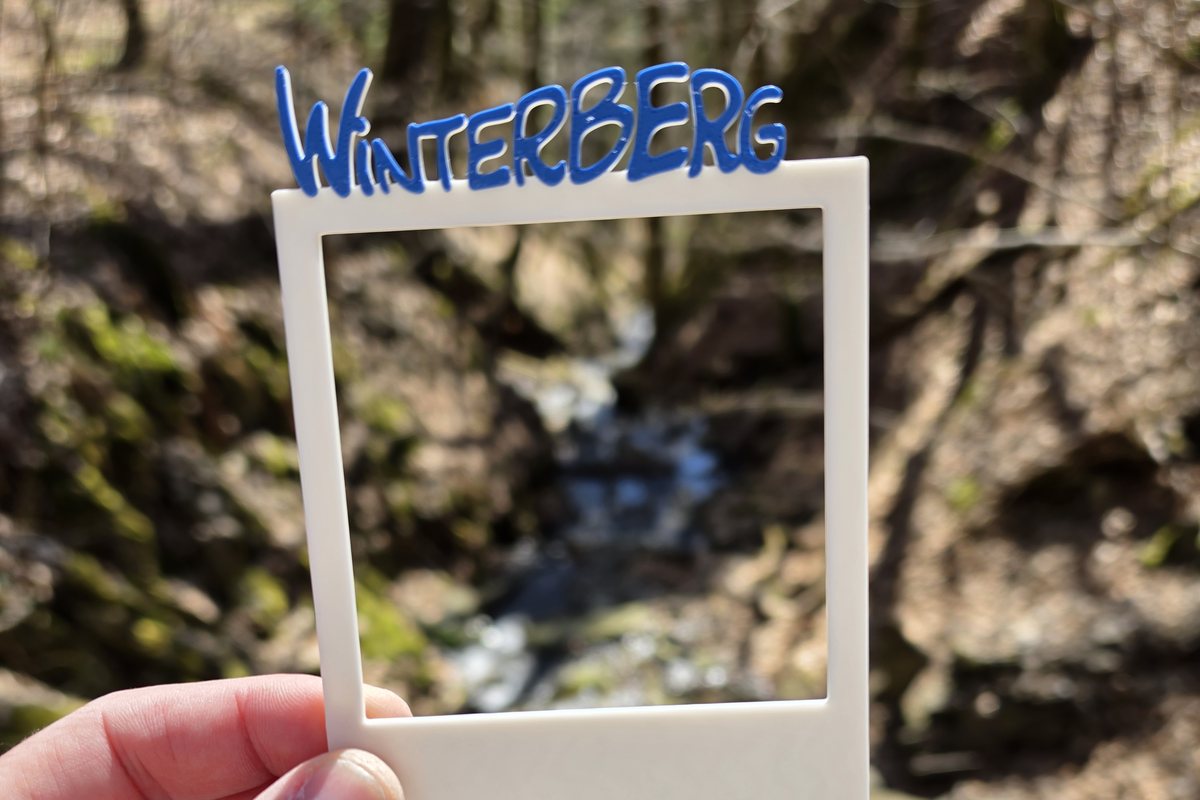 Hand haelt einen selbst gedruckten Fotorahmen mit dem Schriftzug Winterberg vor einen Wasserfall am Schluchten- und Brückenpfad
