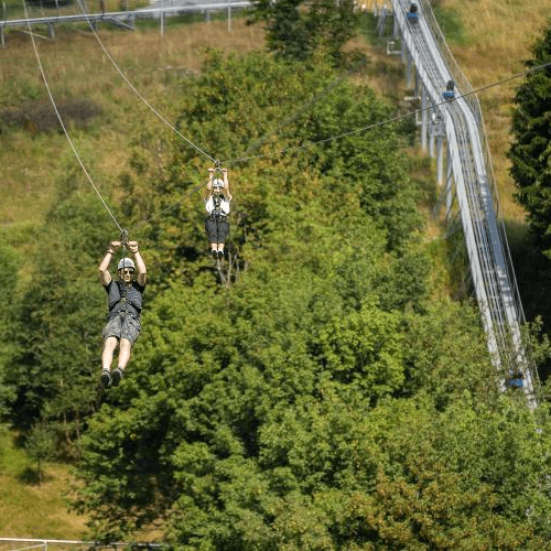 Zwei Personen fliegen mit der Zipline in Winterberg, im Hintergrund die Sommerrodelbahn und grüne Landschaft